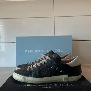 Philippe model skor - Säljer ett par svarta Philippe Model sneakers med vita snören och vit sula. Skorna har gråa och röda detaljer. Klassisk låg modell med rund tå och platt sula. Perfekta för dig som gillar stilrena skor☑️Finns såklart vissa defekter till exempel lite slitna skosnören men sen ska det inte vara ett problem. Storlek 43 men passar även 42