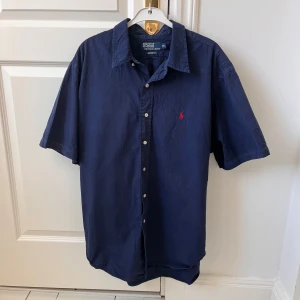 Mörkblå kortärmad skjorta från Polo Ralph Lauren - Säljer en stilren mörkblå skjorta från Polo Ralph Lauren med korta ärmar och klassisk krage. Skjortan har en broderad röd logotyp på bröstet och knappar framtill. Perfekt för en avslappnad men ändå elegant look. Ser lite hängig ut men är i väldigt bra skick