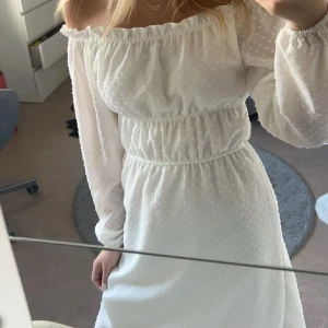 Vit offshoulder klänning med lång ärm - Superfin vit klänning med offshoulder och långa ärmar. Klänningen har resår i midjan och runt axlarna för en snygg passform. Tunt, lätt material med diskret prickigt mönster. Perfekt för sommarens alla tillfällen!