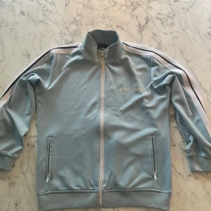 Ljusblå track jacket från Palm Angels - Snygg ljusblå track jacket från Palm Angels med vita och svarta ränder längs ärmarna. Jackan har dragkedja framtill, ståkrage och två fickor med dragkedja. Palm Angels-logga på bröstet.