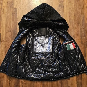 Svart quiltad väst från Philipp Plein - Säljer en svart quiltad väst från Philipp Plein med avtagbar huva och dragkedja framtill. Västen har ett diskret mönster, två fickor med dragkedja och ett stort tryck med texten 'Cowboy' på insidan. Tillverkad i blankt syntetmaterial.