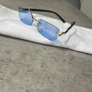 Blå rektangulära solglasögon med gulddetaljer - Snygga solglasögon med rektangulära blåtonade glas och tunna svarta skalmar med gulddetaljer. Glasögonen har en modern och minimalistisk design utan synlig båge runt glasen. Perfekt accessoar för att lyfta din stil.