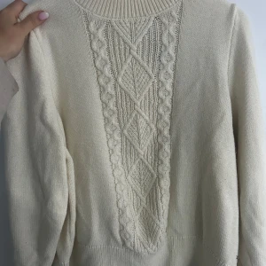 Beige stickad tröja med mönster - Säljer en beige stickad tröja med lång ärm och högt ribbat parti vid halsen. Framsidan har ett snyggt stickat mönster med flätor och diamantformer. Perfekt för dig som gillar klassisk och mysig stil.