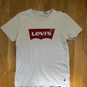 Vit t-shirt med röd Levi's-logga - Säljer en klassisk vit t-shirt från Levi's med den ikoniska röda loggan tryckt på bröstet. T-shirten har rund halsringning och korta ärmar. Perfekt basplagg med snygg och enkel design. Några små defekter och size S