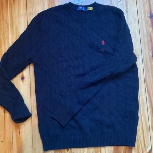 Mörkblå kabelstickad tröja från Polo Ralph Lauren - Säljer en klassisk mörkblå kabelstickad tröja från Polo Ralph Lauren med röd broderad logga på bröstet. Tröjan har rund halsringning och långa ärmar. Perfekt för en stilren och tidlös look.