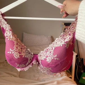 Rosa spets-bh med blommönster - Säljer en snygg rosa bh med vacker vit spets och blommiga detaljer. Bh:n har vadderade kupor och justerbara axelband. Perfekt för dig som vill ha något extra fint under kläderna.