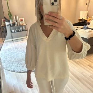 Vit v-ringad blus med trekvartsärm - Säljer en stilren krämvit/ beige blus med v-ringning och trekvartsärmar. Blusen har en lös passform och är tillverkad i ett lätt, mjukt material som ger ett avslappnat intryck. Perfekt att matcha med både jeans och kjol.
