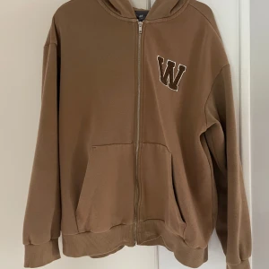 Brun hoodie med dragkedja från H&M - Säljer en brun hoodie från H&M i relaxed fit med dragkedja och huva. Tröjan har två fickor framtill och ett broderat W på bröstet. Perfekt för en avslappnad stil.