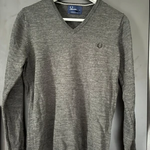 Merinoulls tröja från Fred Perry - Säljer en klassisk grå v-ringad tröja från Fred Perry i 100% merinoull. Tröjan har långa ärmar, broderad logga på bröstet och ribbade muddar vid ärmslut och nederkant. Perfekt för en stilren och enkel look! Har två lagningar, en i mudden och en i nacken.