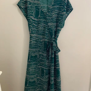 Grön mönstrad omlottklänning från Monki - Säljer en grön långklänning från Monki med vitt abstrakt mönster. Klänningen har omlottmodell med knyt i midjan och korta ärmar. Perfekt för dig som gillar en avslappnad men ändå snygg stil.