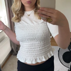 Vit topp - Säljer en söt vit smockad topp med volangdetaljer i nederkant. Toppen har korta ärmar och en tight passform som ger en fin siluett. Perfekt för en somrig look! 