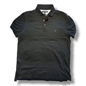 Svart pikétröja från Tommy Hilfiger - Fet polo: Snygg svart pikétröja från Tommy Hilfiger i slim fit. Tröjan har en klassisk krage med knappar och det ikoniska logotypmärket på bröstet. Perfekt för en stilren look.