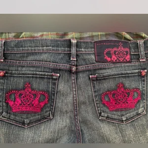 Blå jeans med rosa broderi från Rock & Republic - Snygga blå jeans från Rock & Republic med unika rosa broderade kronor på bakfickorna. Jeansen har låg midja och en bootcut-stil som ger en avslappnad look. De rosa sömmarna och knapparna ger en extra touch av stil. Lånade av tjejen jag köpte av, köpta på depop 🤍           Midja: 34,5 cm rakt över.                                                   Innerbenslängd: 91 cm