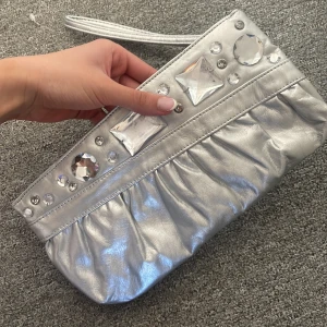 Silverfärgad kuvertväska - Snygg silverfärgad kuvertväska. Väskan har en dragkedja upptill och en praktisk handledsrem. Perfekt för att lägga till lite bling till din outfit!