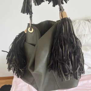 Olivgrön axelväska från River Island - Snygg olivgrön axelväska från River Island med fransar och dragkedja. Väskan har en stilren design med dekorativa detaljer och praktiska fack. Är vintege, hör av dig vid frågor osv❤️