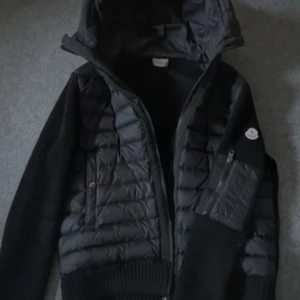 Svart jacka med huva från Moncler - Snygg svart jacka från Moncler med huva och dragkedja. Jackan har quiltade detaljer och en praktisk ficka med dragkedja på ärmen. Perfekt för kyligare dagar.