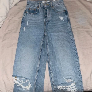 Blå jeans med slitningar från Pull & Bear - Snygga blå jeans med hög midja och slitningar på knäna. De har en klassisk femficksdesign och en knappgylf. Perfekta för en avslappnad stil med en modern twist.