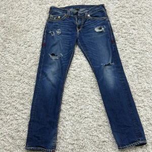 Blå jeans från True Religion - Snygga blå jeans från True Religion med slitna detaljer och kontrastsömmar. Modellen är 'Geno' med en relaxed slim passform. Klassiska femficksdesign och broderad logga på bakfickorna ger en cool look. Använda fåtal gånger (köpta secondhand). En del av sömmarna är lösa eller borta men dom var köpta så. Pris kan diskuteras