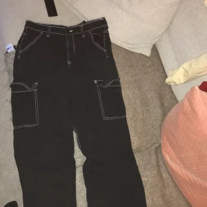 Svarta cargobyxor med vita sömmar - Snygga svarta cargobyxor med kontrasterande vita sömmar. Byxorna har flera praktiska fickor och en loose passform som ger en avslappnad stil. Perfekta för en cool och bekväm look.