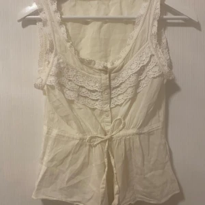 Vit sommar spets volangen ärmlös blus linne med spetsdetaljer - Säljer en söt vit blus med spetsdetaljer och knytband i midjan. Blusen är ärmlös och har en rundad halsringning. Perfekt för en somrig look!
