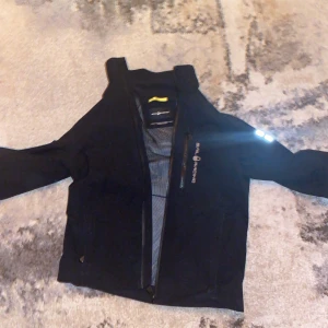 Svart jacka från Sail Racing - Snygg svart jacka från Sail Racing med dragkedja och reflekterande detaljer på ärmarna. Perfekt för utomhusaktiviteter med sin sportiga look och funktionella design.