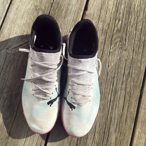 Vita och rosa fotbollsskor från Puma - Snygga fotbollsskor från Puma i vitt med rosa sula och svarta detaljer. Skorna har snörning och en stilren design med Puma-loggan på sidan. Perfekta för inomhusspel med en greppvänlig sula. Som nya! Använt några gånger 