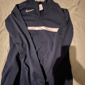 Mörkblå långärmad tröja från Nike - Säljer en mörkblå långärmad tröja från Nike med dragkedja vid halsen och vit rand över bröstet. Tröjan är tillverkad i Dri-FIT material för optimal komfort. Perfekt för träning eller vardagsbruk.