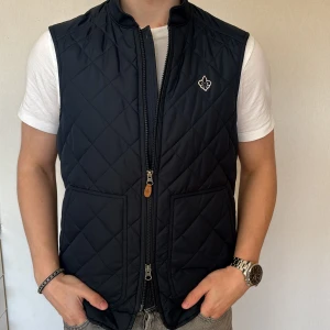 Morris väst - Säljer nu denna Morris väst som jag säljer pga den inte kommit till användning. Snygg svart quiltad väst med dragkedja och två fickor framtill. Västen har en stilren design med en liten broderad detalj på bröstet. Perfekt för lager-på-lager under kyligare dagar. Priset kan diskuteras. Hör av er vid funderingar.