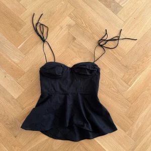 Svart korsettopp från Zara - Snygg svart korsettopp från Zara med smala axelband och dragkedja i sidan. Toppen har en figurnära passform och en lätt utsvängd nederdel. Perfekt för en stilren look. Super fin, har kortat ner banden lite då dom var väldigt långa från början. 