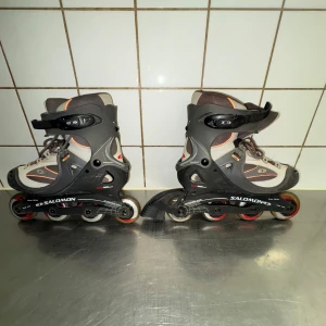 Grå inlines från Salomon - Säljer ett par grå inlines från Salomon med snörning och justerbara spännen. De har en robust design med röd och svart detaljering. Perfekta för en aktiv livsstil och roliga utomhusaktiviteter.