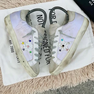 Sneakers med stjärndetaljer - Snygga sneakers med en vit och ljuslila färgkombination. Skorna har en unik stjärndetalj på sidan dekorerad med färgglada stenar. De har grå snörning och en bekväm gummisula.