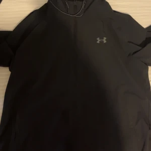 Svart jacka från Under Armour - Säljer en svart jacka från Under Armour med dragkedja och huva. Perfekt för sportiga aktiviteter eller som en lätt ytterjacka. Den har en stilren design med logotyp på bröstet. Säljer den av att den inte passar mig mera. Pris kan även diskuteras  vid snabb intresse. 