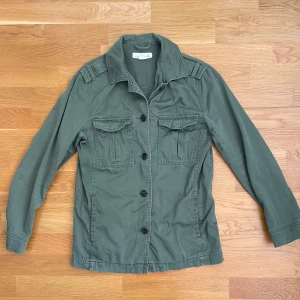 Grön Field jacket - Säljer en stilren grön jacka från logg. Jackan har långa ärmar och knappar framtill samt två bröstfickor med lock. Perfekt för en avslappnad stil.