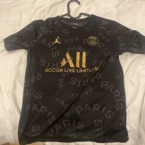 Svart PSG fotbollströja med guldtryck - Snygg svart PSG fotbollströja med guldtryck och texten 'Accor Live Limitless' på framsidan. Tröjan har korta ärmar och ett mönster med ordet 'Paris' över hela tyget. Nummer 7 i guld på baksidan med 'Visit Rwanda' ovanför. Perfekt för fotbollsfans!