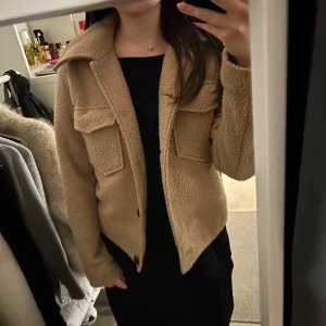 Beige teddyjacka med knappar - Mysig beige teddyjacka med knappar framtill och stora fickor på bröstet. Perfekt till våren! 