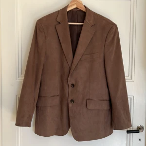 Brun blazer faux suede - Snygg brun kavaj i mockaimitation. Från Dressmann i storlek 48. Kavajen har två knappar framtill och fyra dekorativa knappar vid ärmsluten. Perfekt för en stilren look.