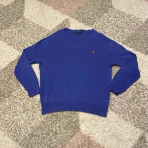 Blå tröja från Polo Ralph Lauren - Säljer en snygg blå tröja från Polo Ralph Lauren med v-ringning och det klassiska logotypbroderiet i orange på bröstet. Tröjan har långa ärmar och är perfekt för en stilren look. Passar bra till både jeans och chinos. Storlek L