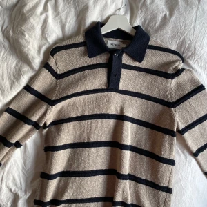 Randig pikétröja från East West - Säljer en stilren randig pikétröja från East West i beige och svart. Tröjan har en klassisk krage och korta ärmar, perfekt för en avslappnad look. Passar bra till både jeans och chinos. Använd en gång, tvättad en gång 