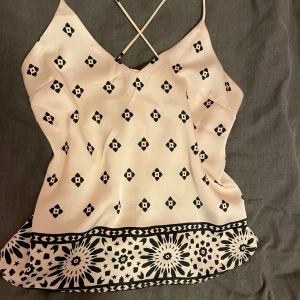 Beige och svart mönstrad linnetopp - Snygg beige och svart mönstrad linnetopp med tunna axelband och korsad rygg. Perfekt för varma dagar eller som ett lager under en kavaj. Den har ett unikt geometriskt mönster och en bredare kant med blommönster längst ner.