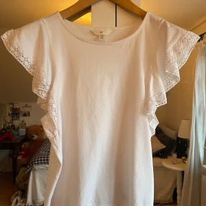 Vit blus från H&M - Säljer en söt vit blus från H&M med korta ärmar och spetsdetaljer. Perfekt för en somrig look med sina volanger på ärmarna. Blusen har en rund halsringning och är gjord i ett mjukt material.