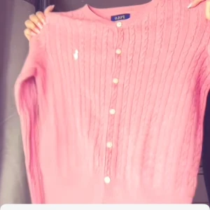 Rosa kofta från Ralph Lauren - Säljer en söt rosa kofta från Ralph Lauren med kabelstickat mönster och knappar framtill. Den har långa ärmar och en klassisk design med en liten logga på bröstet. Perfekt för att lägga till en färgklick i garderoben!