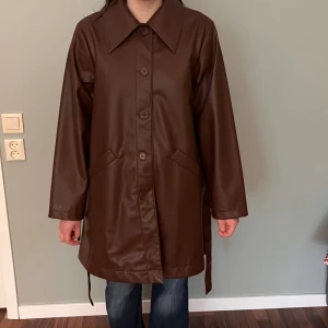 Brun skinnjacka med bälte - Snygg brun skinnjacka med knappar och bälte i midjan. Jackan har en klassisk krage och långa ärmar. Perfekt för en stilren look.