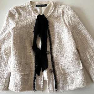 Vit jacka från Zara Woman - Klassisk prêt-à-porter kavaj / jacket, fina dekorativa detaljer. Passar bra till office wear eller casual jeans. En elegant vit jacka. Jackan har en chic textur och är dekorerad med en svart rosett framtill. Den har långa ärmar och knappar för stängning. Perfekt för en stilren look! 