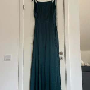 Mörkgrön långklänning - Elegant mörkgrön långklänning i satin med omlott-design och smala axelband. Perfekt för en stilfull kväll med en vacker drapering framtill. Klänningen har även en spets detalj i ryggen och slits💕