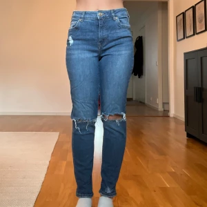 Blåa mom jeans med slitningar - Snygga blåa mom jeans med slitningar vid knäna. De har en klassisk femficksdesign och är högmidjade för en bekväm känsla. Perfekta för en avslappnad stil.