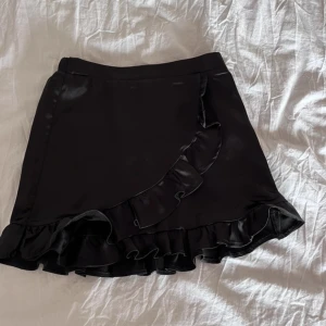 Svart kjol från Gina Tricot - Snygg svart kjol från Gina Tricot med volangdetaljer och omlott-design. Använd max fem gånger. Från Young avledningen i storlek 146/152, passar mig som i vanliga fall har XXS-XS