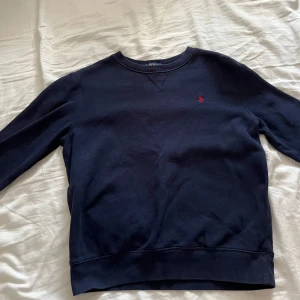 Mörkblå tröja från Polo Ralph Lauren - Säljer en stilren mörkblå tröja från Polo Ralph Lauren med det klassiska röda logotypbroderiet på bröstet. Tröjan har långa ärmar och rund halsringning, perfekt för en avslappnad look.