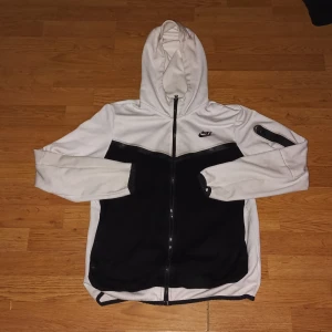 Vit och svart hoodie från Nike - Säljer en snygg vit och svart hoodie från Nike med dragkedja och huva. Jackan har en cool design med svarta paneler och Nike-logga på bröstet. Perfekt för en sportig look eller chill vardagsstil.