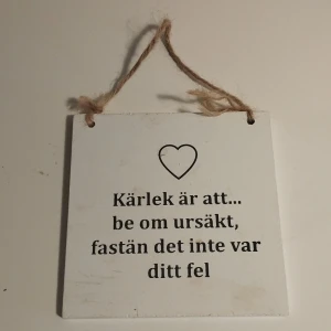 Vit skylt med text från FORM Living - Charmig vit skylt i trä från FORM Living med texten 'Kärlek är att... be om ursäkt, fastän det inte var ditt fel'. Skylten har ett litet hjärta ovanför texten och hängs upp med ett snöre. Perfekt för att sprida lite kärlek och visdom i hemmet.  Storlek 10x10 cm.