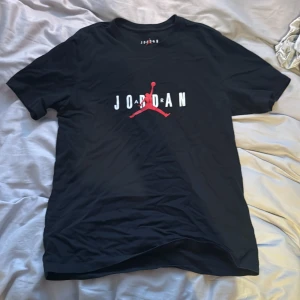 Svart t-shirt från Jordan - Snygg svart t-shirt från Jordan med det ikoniska Jumpman-logotypen i rött och texten 'JORDAN' i vitt på både fram- och baksidan. Perfekt för en sportig och avslappnad stil.